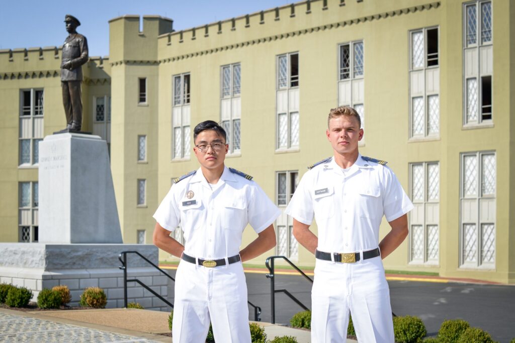 Two male cadets pose in white class uniform in front of the Marshall statue.