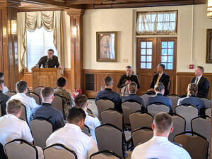 Lt. Col. Mark Boonshoft stands at a podium with judges seated to his left.