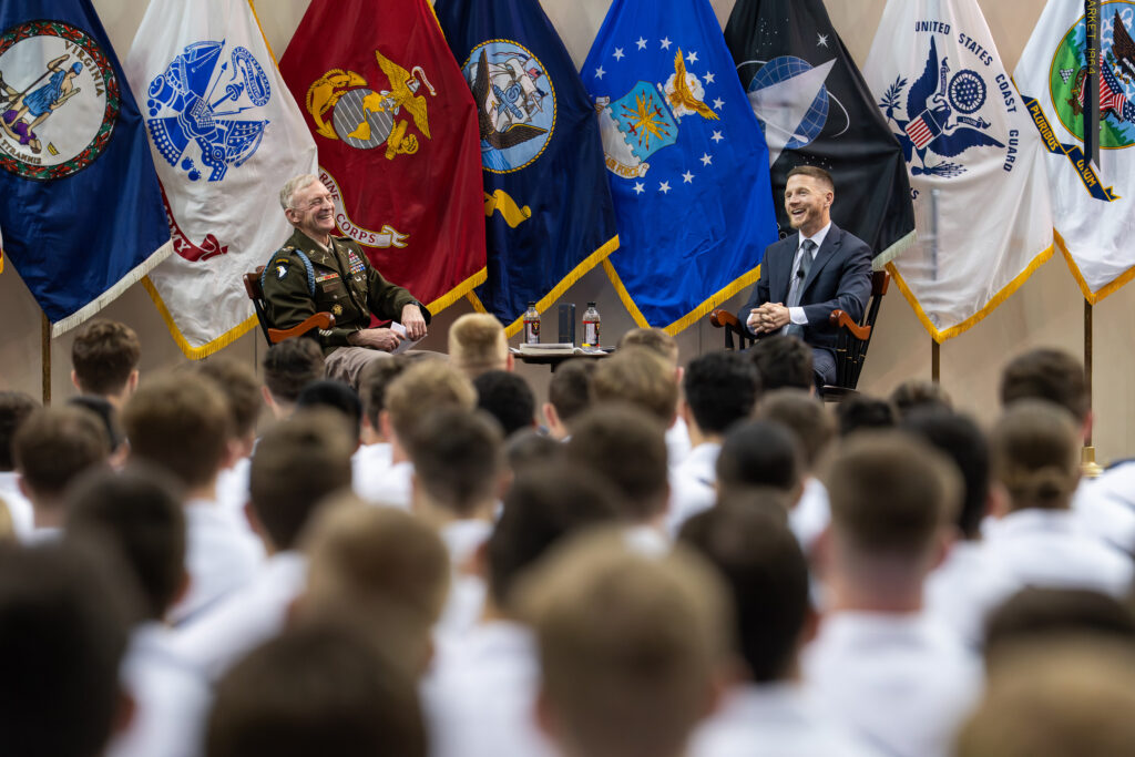 CLE director and Kyle Carpenter sit on a stage in fronts of military flags while VMI cadets listen.